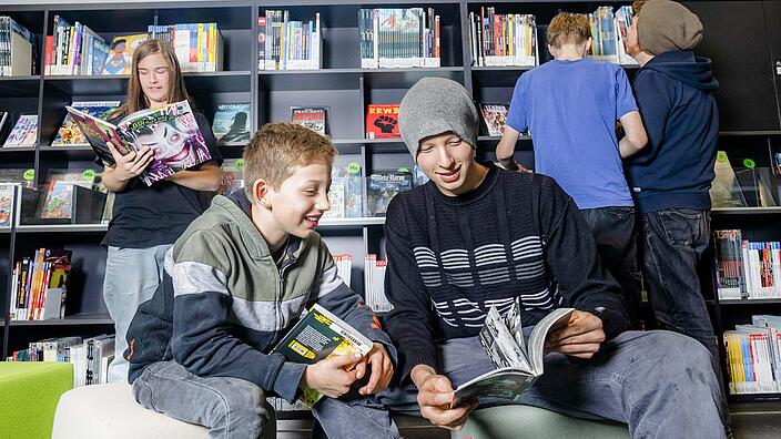 Junge und Jugendliche stehen oder sitzen in Zeitschriften-Ecke der Bibliothek mit Zeitschriften