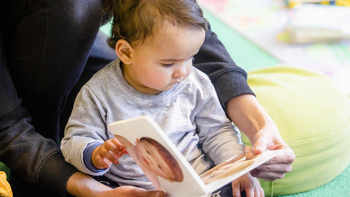 Kleinkind sitzt auf Boden in Bibliothek und betrachtet ein Babybuch, das Erwachsener festhält