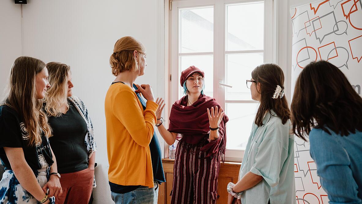 Sechs Personen stehen in einem Raum. Eine von ihnen steht vor einem Fenster und spricht, die anderen hören ihr zugewandt zu.