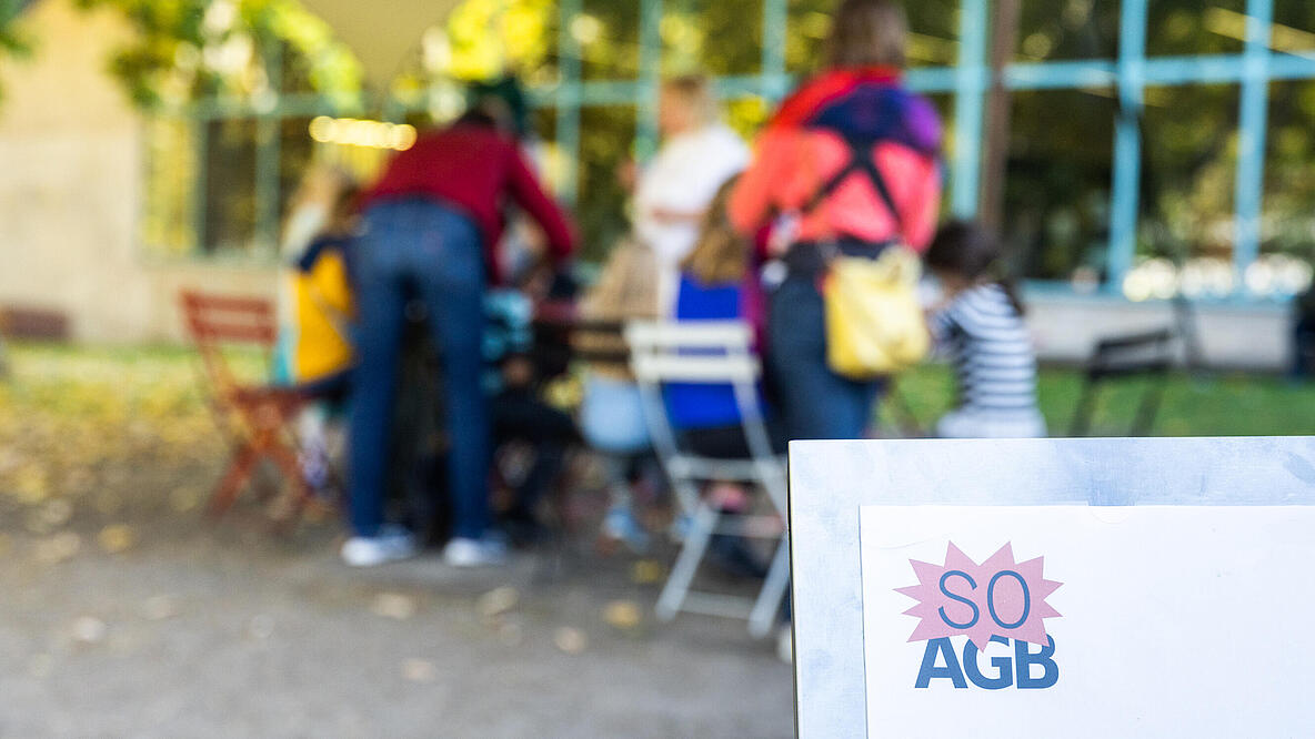 Forschergarten Programmschild im Vordergrund vor verschwommener Tischgruppe mit sitzenden und stehenden Menschen