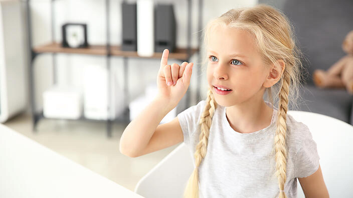 Ein blondes Mädchen mit zwei geflochtenen Zöpfen macht mit der Hand eine Gebärde.