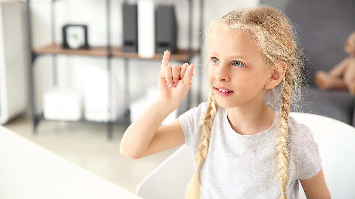 Ein blondes Mädchen mit zwei geflochtenen Zöpfen macht mit der Hand eine Gebärde.