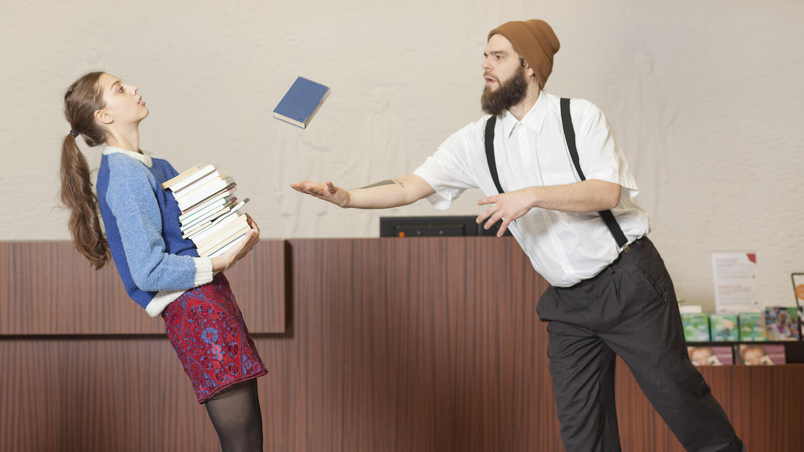 Frau mit Bücherturm auf Arm kippt nach hinten, während Mann sich zu ihr und fallendem Buch lehnt