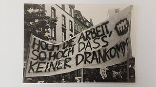 Schwarz-weiß-Bild eines Demonstrationsumzugs mit einem Banner mit der Aufschrift "Hoch die Arbeit, so hoch dass keiner drankommt"