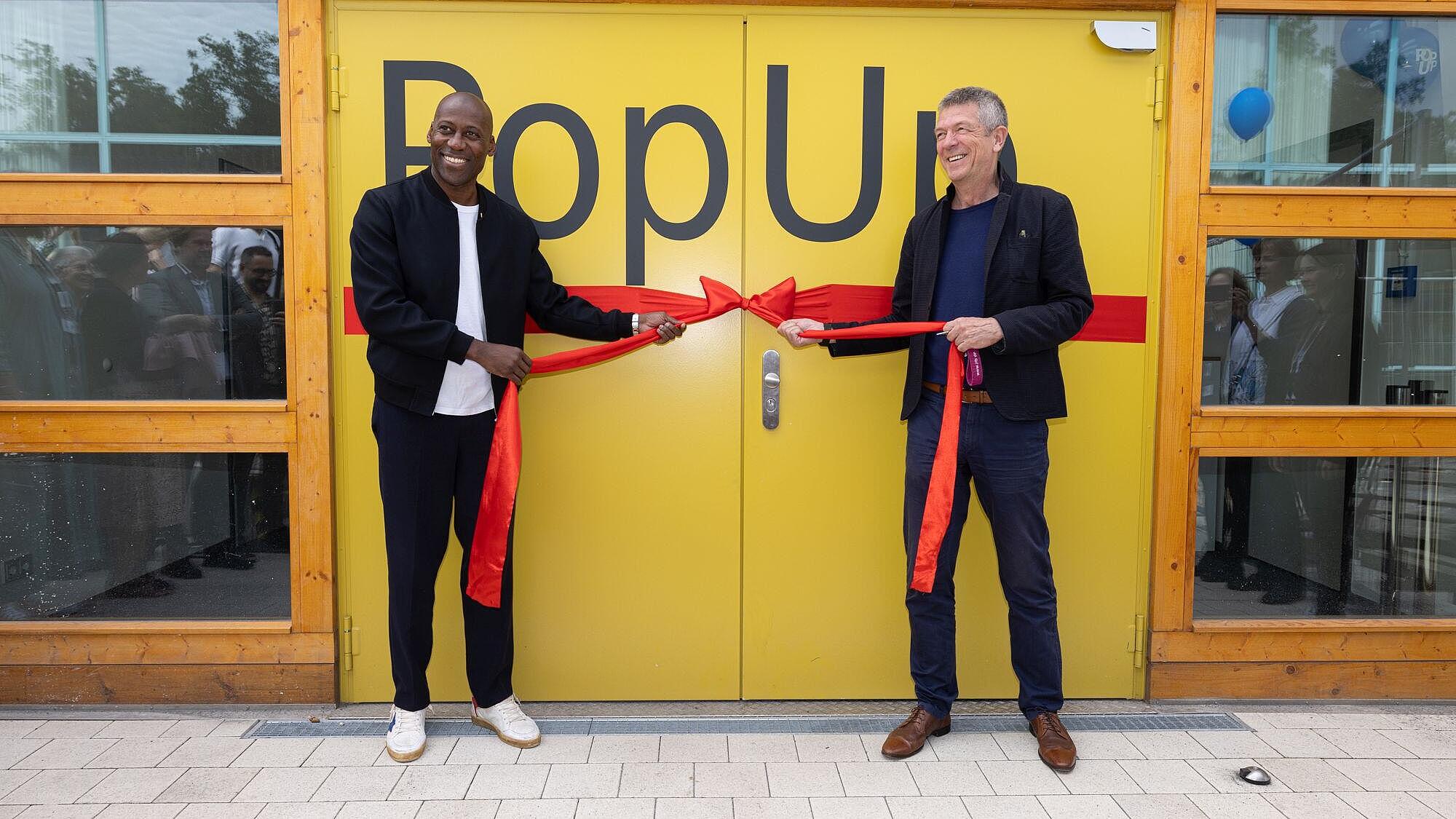 Foto von Kultursenator Joe Chialo und Generaldirektor der ZLB Volker Heller vor der Eingangstür zum PopUp-Gebäude. In den Händen halten sie eine große rote Schleife zur Eröffnung.