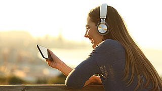 Junge Frau mit Kopfhörern schaut Videos auf Smartphone in der Sonne